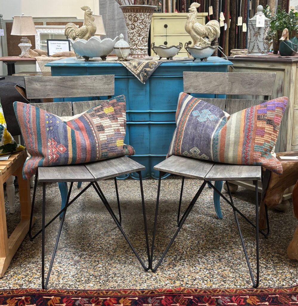 Two rustic grey and black loft chairs with colorful kilim pillows are displayed in a cozy furniture store setting. The chairs feature sleek metal legs and wooden backs, paired with vibrant, patterned pillows in various colors. The background includes a turquoise sideboard, decorative items, and other rustic furniture pieces, creating an inviting atmosphere.